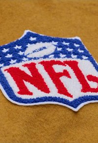NFL-Logoaufnäher mit roten Buchstaben auf weißem Hintergrund, umgeben von blauen Sternakzenten, auf einem beigefarbenen, strukturierten Stoffuntergrund.