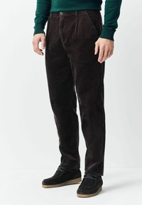 Man wearing dark brown corduroy pants, black casual shoes, and a dark green long-sleeve shirt, standing on a white background.