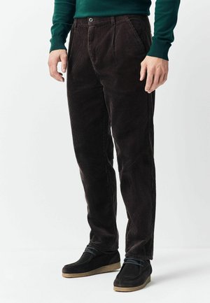 Mann trägt dunkelbraune Cordhosen, schwarze Freizeit-Schuhe und ein dunkelgrünes Langarmhemd, steht vor einem weißen Hintergrund.