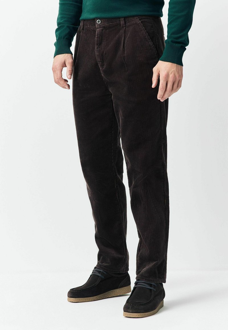 Man wearing dark brown corduroy pants, black casual shoes, and a dark green long-sleeve shirt, standing on a white background.