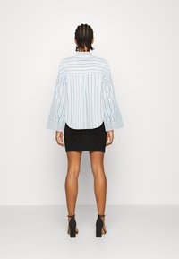 Hellblaue gestreifte Oversize-Bluse mit Glockenärmeln, kombiniert mit einem schwarzen, figurnahen Rock und schwarzen Sandalen mit Absatz.