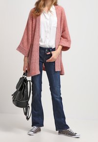 Rosa, öppen cardigan över en vit skjorta, tillsammans med mörkblå utsvängda jeans och grå sneakers. Svart ryggsäck med en klaffdetalj.