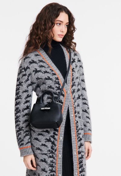 Schwarze strukturierte Handtasche mit einem oberen Griff und Schulterriemen, ausgestattet mit einer glatten Textur und silbernem Zubehör. Getragen mit einer grauen gemusterten Strickjacke.