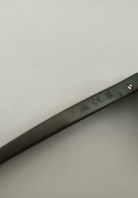 Montura de gafas de sol de plástico negro con una textura suave, que presenta acentos metálicos y una marca en relieve en la patilla.