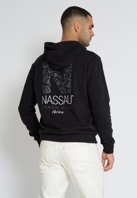 Musta huppari, jossa on suuri, teksturoidulla graafikuviointi takana, jossa lukee "NASSAU BEACH CLUB" ja "Ibiza", yhdistettynä valkoisiin farkkuihin.