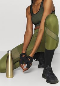 Guanti sportivi neri con ritagli sulle dita, leggings verde oliva con pannelli in rete, scarpe sportive nere e una bottiglia d'acqua in oro metallico.