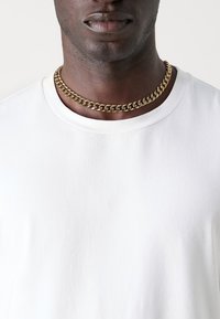 Goldene Kettennacklace mit miteinander verbundenen Gliedern, getragen über einem schlichten weißen Baumwoll-T-Shirt mit rundem Ausschnitt, sichtbar strukturiert und mit glatter Oberfläche.