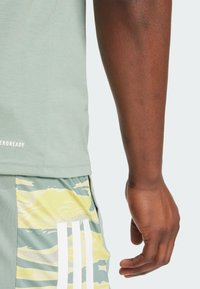 Kortärmad ljusgrön skjorta med rak fåll och mjuk textur, ihop med mönstrade shorts i gul och grön kamouflage med tre vita ränder.
