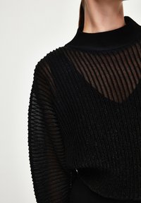 Svart ribbad tröja med hög krage, genomskinliga texturerade paneler och subtil glittrande detalj, som framhäver en kombination av ogenomskinliga och genomskinliga material.