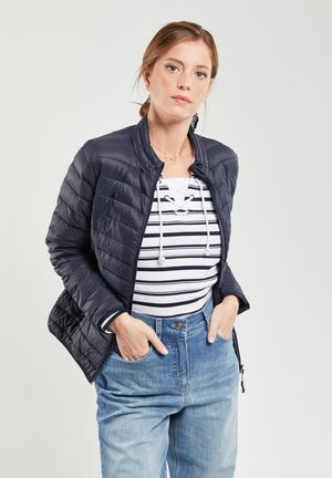 Blouson aviateur matelassé bleu marine avec col montant, fermeture éclair et poignets élastiqués. Haut rayé blanc et bleu en dessous ; jean bleu clair.