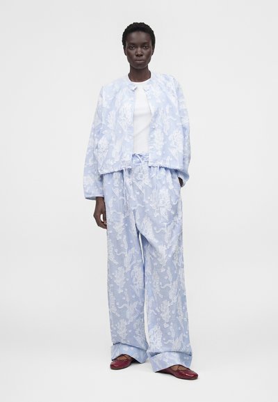 Γυναίκα που φοράει ανοιχτό μπλε σακάκι με λουλουδάτο σχέδιο και αντίστοιχο παντελόνι με φαρδιά πόδια, λευκή μπλούζα και μπορντό πλατφόρμες, στέκεται σε λευκό φόντο.