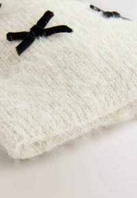 Accessoire tricoté blanc avec une texture pelucheuse, orné de nœuds en velours noir, présentant un design doux et délicat avec une légère brillance.