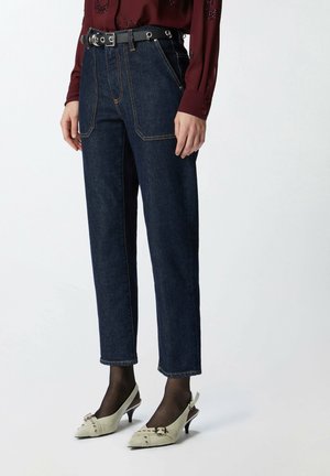 Femme portant un jean taille haute bleu foncé avec poches avant, ceinture noire, blouse bordeaux à manches longues, collants noirs transparents et chaussures blanches à talons.