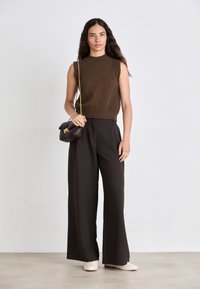 Braun ärmelloses Strickoberteil kombiniert mit hochtaillierten, weit geschnittenen dunklen Hosen. Die Accessoires umfassen eine kleine bordeauxfarbene Handtasche und cremefarbene Loafers.