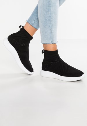 Füße in schwarzen Calvin Klein Sock-Sneakern mit weißen Sohlen und hellblauen Jeans mit ausgefranstem Saum, stehend auf einem glatten weißen Boden.