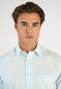 Lichtblauw overhemd met knopen en een zak, met geborduurd "L." Gemaakt van een gladde stof met een puntkraag en ivoren knopen.