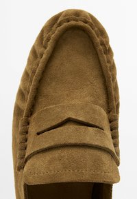 Brązowy zamszowy mokasyn z przeszyciami w stylu mokasynowym i paseczkiem na górze, widziany z góry na białym tle.