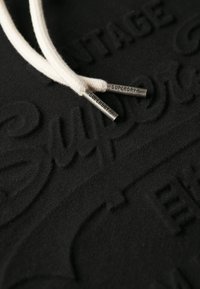 Sudadera negra con un diseño de logotipo texturizado. Cuerdas de algodón en color crema con puntas metálicas grabadas con "SUPERDRY".