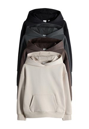 Dziecko z włosami do ramion opierające się o ścianę, ubrane w beżową bluzę z kapturem, szerokie niebieskie dżinsy oraz brązowe buty wsuwane, uśmiechające się i patrzące w górę.