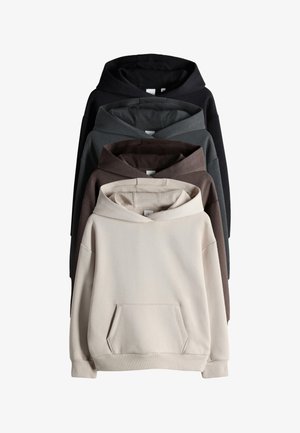 Dziecko z włosami do ramion opierające się o ścianę, ubrane w beżową bluzę z kapturem, szerokie niebieskie dżinsy oraz brązowe buty wsuwane, uśmiechające się i patrzące w górę.