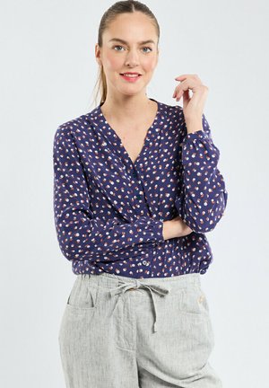 Blouse navy avec un petit motif floral, col en V et détail de boutons. Manches longues avec poignets froncés. Associée à un pantalon en lin gris clair.