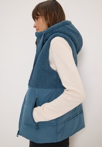 Blauer Pufferweste mit einem oberen Bereich aus Fleece und Kapuze, ausgestattet mit einer Frontreißverschlusstasche, weicher Textur und einem kontrastierenden Materialmix.