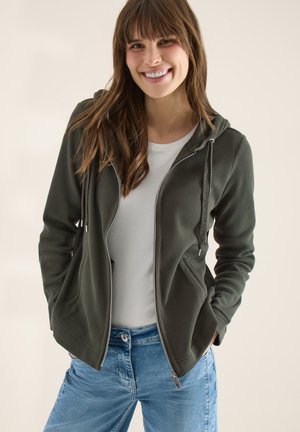 Femme souriante aux cheveux bruns portant un sweat à capuche vert zippé sur un t-shirt blanc et un jean bleu, mains dans les poches, fond neutre.