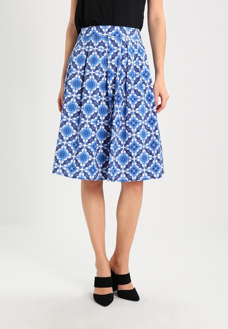 Femme portant une jupe à motifs géométriques bleus et blancs mi-longue, un haut sans manches noir et des talons carrés noirs à bout pointu.