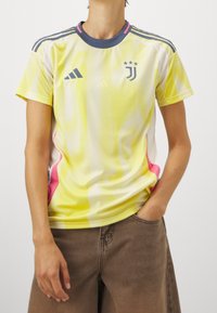 Personne portant un maillot de football jaune de la Juventus avec un col bleu marine et des accents roses sur les côtés, associé à un pantalon marron.