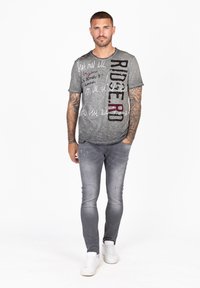 Camiseta gris de manga corta con tejido texturizado, que presenta una impresión en negrita "RIDERD" en negro y marrón, combinada con jeans gris desgastados de corte ajustado y zapatillas blancas.