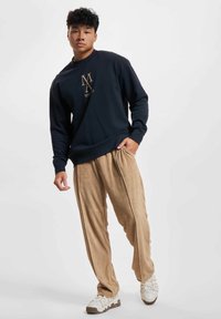 Young man wearing a navy sweatshirt with beige text, beige pants, and white sneakers, standing with one hand in his pocket.