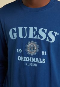 Blaues Langarm-T-Shirt aus Baumwolle mit großem, hellblauem "GUESS"-Logo, das ein dekoratives Badge und darunter stehenden Text enthält.