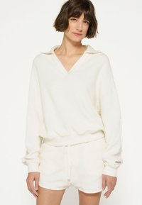 Felpa bianca e comoda con scollo a V, maniche lunghe e polsini a costine, abbinata a pantaloni corti bianchi coordinati. Tessuto morbido e texturizzato con un taglio rilassato.