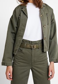 Oliivinvihreä lyhyt takki etupainikkeilla ja vetoketjullisilla taskuilla, yhdistettynä samansävyisiin korkeavyötäröisiin housuihin ja valkoiseen t-paitaan.