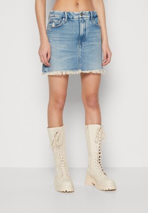Model draagt een lichtblauwe spijkerbroek minirok met rafelige zoom, gecombineerd met crèmekleurige veterlaarzen tot aan de knie.