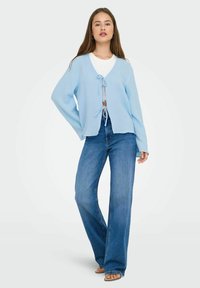 Cardigan azzurro chiaro in maglia texturizzata con una chiusura frontale e design avvolgente, abbinato a jeans blu a vita alta.
