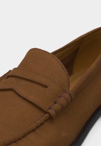 Καφέ καστόρινη μοκασίνι με ραφές μοκασίνου και διακοσμητική λουρίδα σε μαύρη σόλα πάνω σε λευκό φόντο.