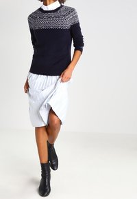 Marineblauer Pullover mit gemustertem Einsatz, kombiniert mit einem hell gestreiften Wickelrock. Schwarze Knöchelschäfte runden den Look ab. Glatte Textur und figurschmeichelnde Passform.