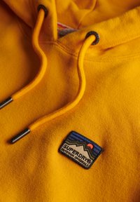 Felgekleurde gele hoodie met een trekkoord, voorzien van zwarte aglets. Inclusief een patch met bergontwerp en "SUPERDRY®" branding. Zachte textuur.