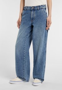 Weite blaue Denim-Jeans, getragen von einer Person, kombiniert mit hellen Turnschuhen, vor einem schlichten weißen Hintergrund.
