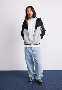 Siva in črna zadrgasta jakna s kapuco, v kombinaciji z belo majico in ohlapnimi svetlo modrimi jeans hlačami. Črno-bela športna obutev z vzorcem.