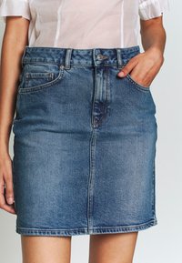 Žena v modré džínové sukni s předními kapsami a knoflíkem, v kombinaci s průhledným bílým topem s krátkým rukávem, ruka v kapse.