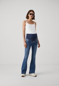 Graviditetsantrekk med en hvit tanktop, mørkeblå over-bump midjebånd og utslåtte blå jeans. Modellen har på grå joggesko og solbriller.