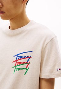 Beige bavlněné tričko s kulatým výstřihem, s barevným textem v modré, červené a zelené barvě v podobě štětcových tahů. Malé logo na levém rukávu.