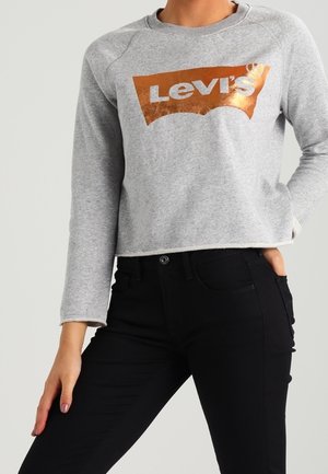 Osoba ubrana w szarą krótką bluzę z długim rękawem z metalicznym pomarańczowym logo Levi's i czarne dopasowane spodnie, pozująca z jedną ręką w kieszeni.