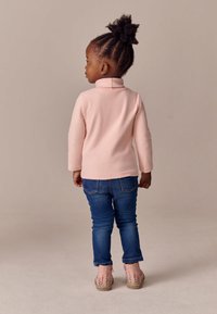Barn iført en pastelrosa fleece-trøje, mørkeblå jeans og lyserøde sko med floral broderi, set bagfra mod en neutral baggrund.