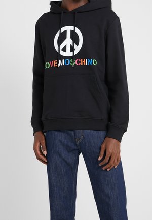 Czarna bluza z kapturem z białym symbolem pokoju i kolorowym napisem "LOVE MOSCHINO", noszona z niebieskimi dżinsami.