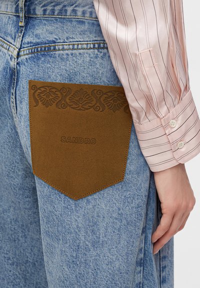 Τζιν από denim με μεγάλη καφέ τσέπη από σουέντ με ανάγλυφα λουλουδάτα μοτίβα και λογότυπο "Sandro", συνδυασμένο με ροζ ριγέ πουκάμισο με μακριά μανίκια.