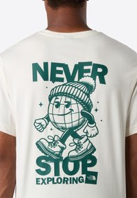 Person iført en hvid T-shirt med et grønt grafisk motiv af en smilende jordklode i støvler og en hue, samt teksten "NEVER STOP EXPLORING."