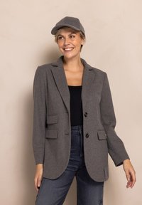 Grauer Blazer aus Wollmischung mit zwei Fronttaschen und zwei schwarzen Knöpfen. Kombiniert mit einer passenden grauen Mütze. Das Modell trägt dunkle Jeans und ein schwarzes Oberteil.